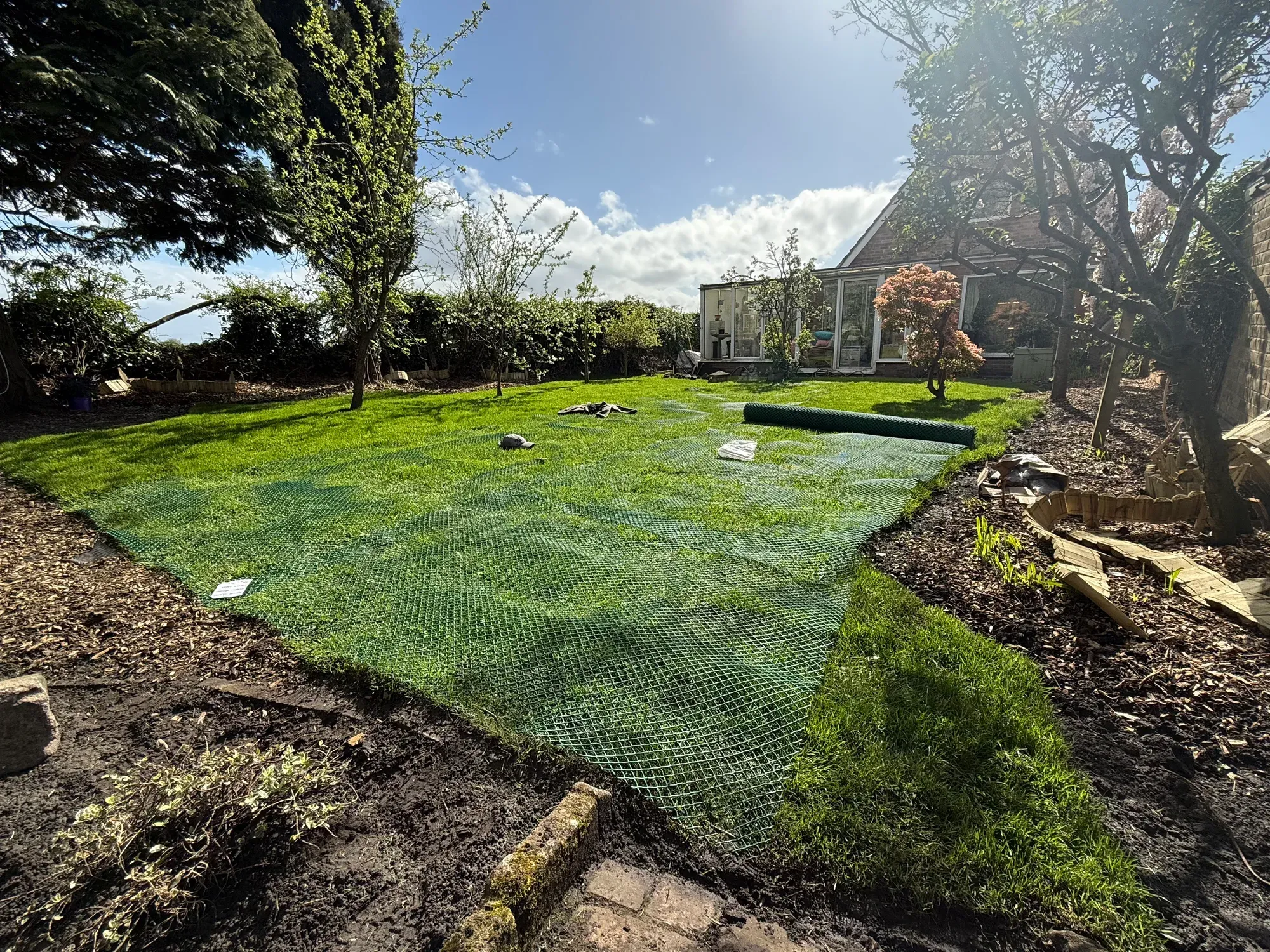 A newly laid lawn under protective green mesh, with mature trees and a greenhouse behind